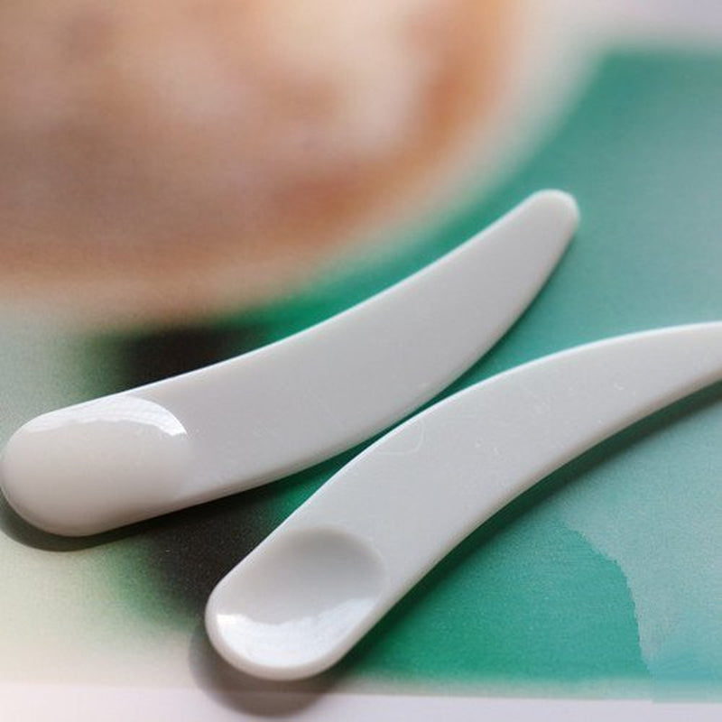 Set of 100 white mini curved cosmetic spoons by AuraLab for makeup and mask application, made of durable plastic.